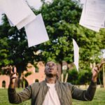 Resume - Overjoyed African American graduate tossing copies of resumes in air after learning news about successfully getting job while sitting in green park with laptop